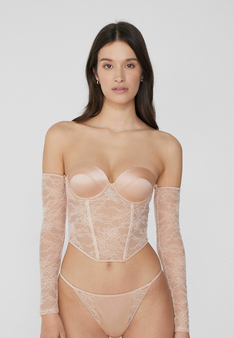 Conjunto de corsé sin tirantes y tanga en color nude; presenta copas de satén y un cuerpo de encaje transparente con largos mangas de encaje, patrones florales y un diseño ajustado.