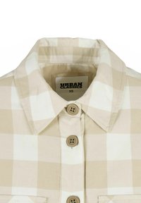 Chemise à carreaux beige avec un col boutonné, présentant de grands motifs carrés, quatre boutons sur le devant et une étiquette indiquant "Urban Classics XS".