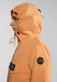 Oranje nylon jack met capuchon, voorzien van elastische koordjes, een ritssluiting en een patch op de linkermouw. Glad textuur en lichtgewicht ontwerp.
