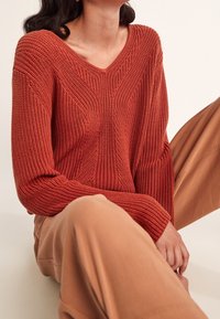 Sweter w kolorze rdzy, dziany, z dekoltem w kształcie litery V, z prążkowanym wzorem i geometrycznym wzorem, w połączeniu z beżowymi spodniami o szerokich nogawkach.