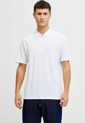 Joven con una camiseta polo blanca de manga corta con cremallera frontal, combinada con jeans azul oscuro, de pie frente a un fondo liso.