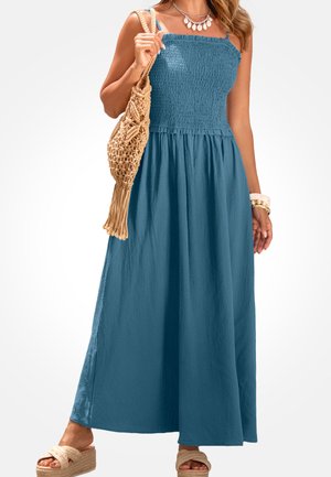 Vrouw draagt een blauwe strapless maxi-jurk met geplooide details, een beige geweven handtas, een gelaagde schelpenketting, armbanden en beige platformsandalen.