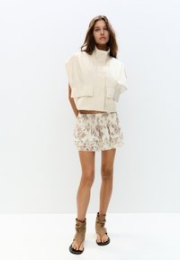 Giacca cropped beige chiaro con maniche corte e due tasche frontali, abbinata a una mini gonna a strati con stampa floreale e sandali con tacco color sabbia.