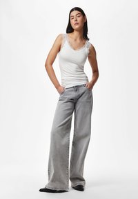 Mujer con una camiseta sin mangas blanca con borde de encaje y pantalones vaqueros grises de pierna ancha, de pie con las manos en los bolsillos sobre un fondo blanco.