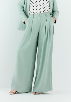 Frau trägt mintgrüne Hosen mit weitem Bein und passenden Blazer, kombiniert mit weißer gepunkteter Bluse und beigen Sandalen mit offenen Zehen.