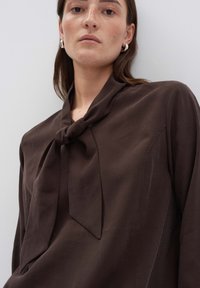 Donna che indossa un blouse marrone scuro con un dettaglio a fiocco, orecchini a cerchio in argento, e un'espressione neutra su uno sfondo semplice.
