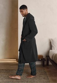Rochie de baie gri cărbune cu glugă, buzunare laterale și un cordon, purtată peste pantaloni în carouri verzi și bleumarin, pe un fundal neutru.