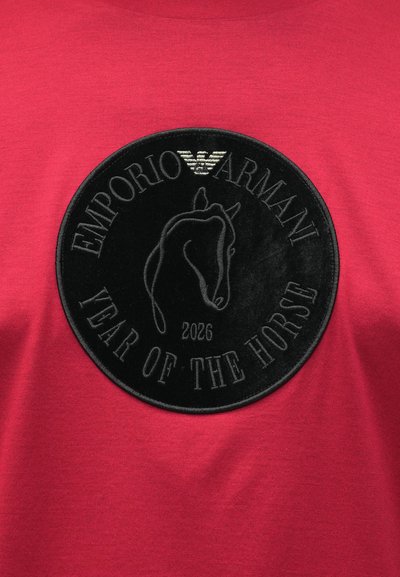 Rdeča bombažna majica z izrazito črno krožno značko z motivom konja in reliefnim napisom »Emporio Armani Leto konja 2026«.