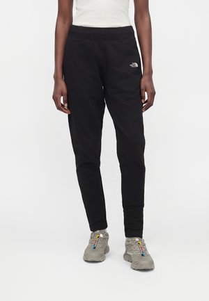 Person iført sorte North Face joggerbukser og grå sneakers med røde, gule og blå accenter på en hvid baggrund.