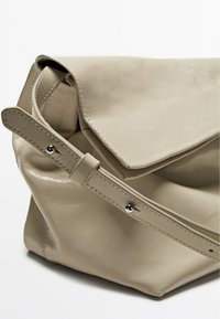 Beige leather bag with a fold-over flap, curved shape, and smooth texture. Features a strap with a silver snap detail.