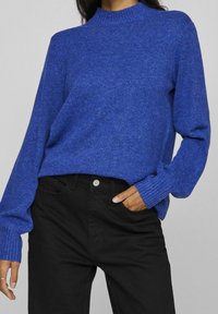 Luxe blauwe gebreide trui met een hoge hals en geribbelde boorden, gecombineerd met zwarte pantalons met een zilveren knoopdetail.