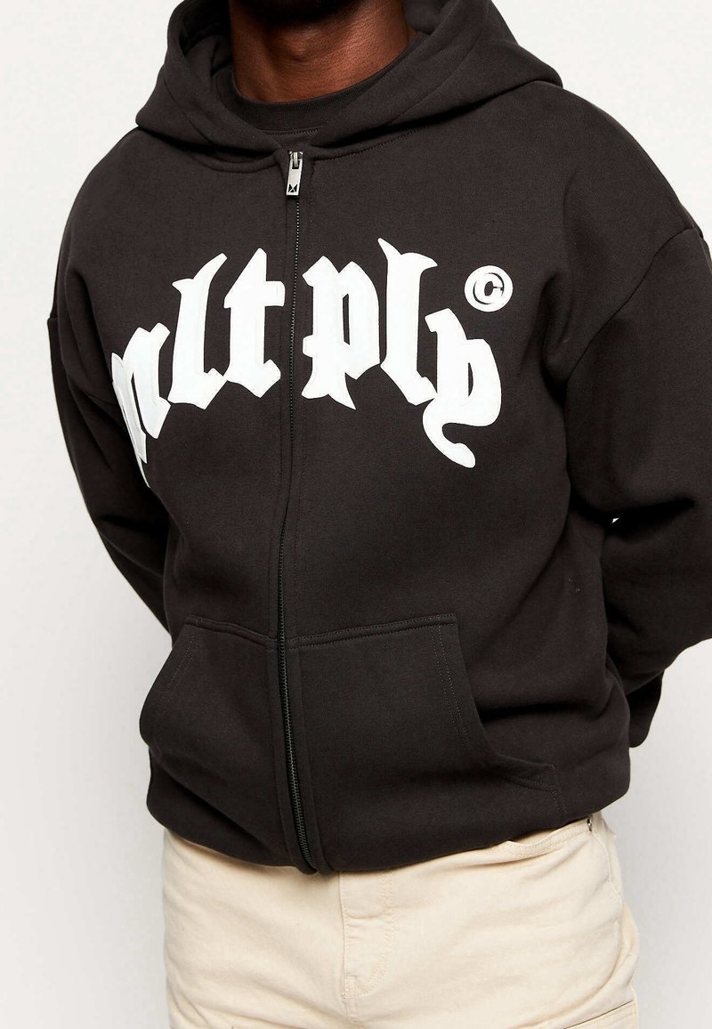 Zwarte hoodie met rits, gemaakt van zacht materiaal, met grote witte letters op de voorkant, kangoeroezakken en een capuchon met trekkoord.