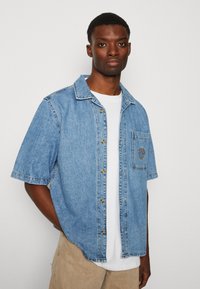 Gaiši zils denima kreklis ar īsām piedurknēm, ar pogu aizdari, kabatas detaļu un apļa logo plāksteri. Ievērojama vaļīga piegriezuma un izliekta apakšmala.