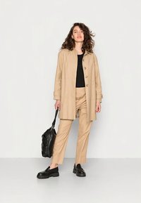 Bež plašč s gumbi, črn zgornji del, bež hlače in črni masivni čevlji. Stiliran z črno torbico, stoječe na nevtralnem ozadju.