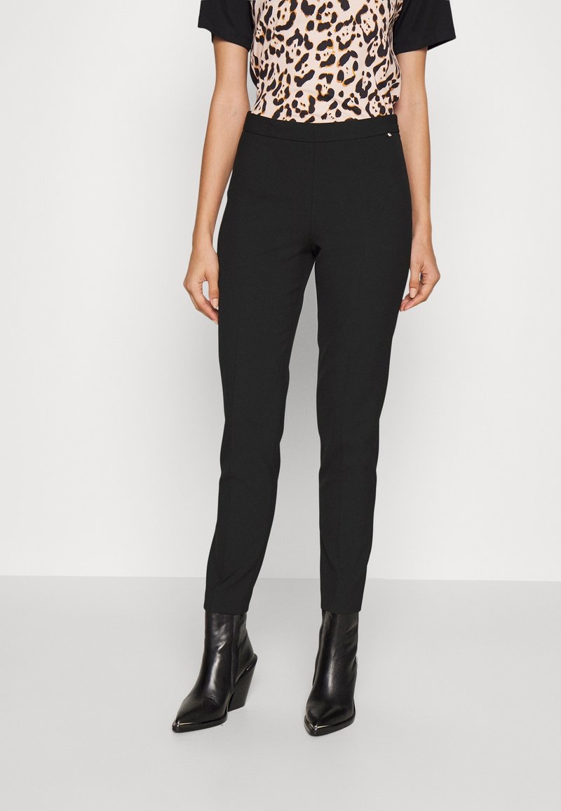 Femme portant un pantalon noir slim, un haut imprimé léopard et des bottines noires à talons, debout devant un fond clair et uni.