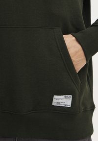 Felpa verde scuro realizzata in tessuto di alta qualità, con una tasca frontale e un'etichetta con istruzioni per la cura cucita sull'orlo.