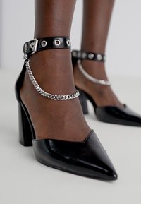 Sorte patentlæder høje hæle med spids tå, ankelrem med metalnitter og kædeaccent. Blockhæl design.