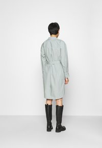 Světle šedé šaty košilového střihu s dlouhými rukávy a páskem v pase, s knoflíkovou zadní stranou, sahající do poloviny stehna, kombinované s černými kozačkami do kolen.