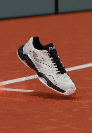 Hvit og svart tennissko med lisser som sparker opp leirstøv på en rød grus tennisbane nær hvite grenselinjer.