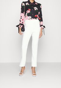 Blusa floreale con motivi neri e rosa, maniche lunghe con fiocchi; pantaloni bianchi su misura; abbinati a sandali a tacco rosa chiaro.