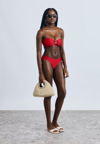 Červené bikiny s bandeau topom s uzlom vpredu, doplnené o zladené nohavičky. Modelka drží béžovú tkanú kabelku a nosí čierne slnečné okuliare.
