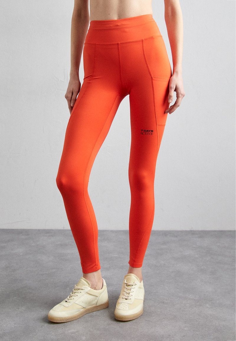 Red high-waisted leggings with a smooth texture, featuring a logo on the lower leg and stitched detailing. Worn with light-colored sneakers.