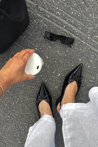 Ballerines en cuir verni noir à bout pointu avec un détail de nœud, associées à un pantalon large gris clair. Un sac noir et des lunettes de soleil se trouvent à proximité.