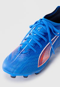Klart blå fotbollssko med snörning, texturerad yta och orange-vita detaljer mot en vit bakgrund.