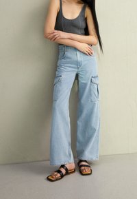 Žena s dlouhými černými vlasy, oblečená v šedém tílku bez rukávů, světlých volných modrých cargo džínách a černých sandálech na širokých popruzích, stojící se založenýma rukama.