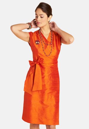 Woman adjusting necklace wearing sleeveless orange wrap dress with large side bow and beaded jewelry against plain background.