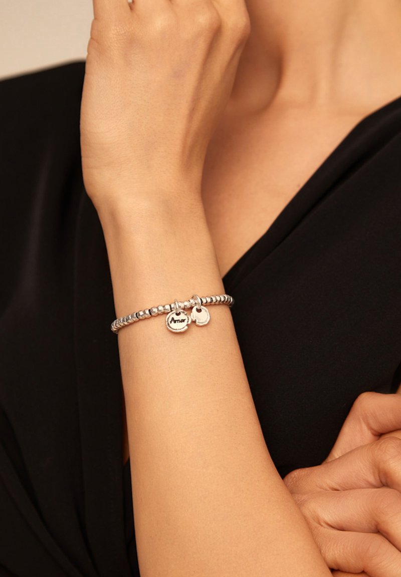 Bracelet à breloques en argent avec des perles rondes, comprenant deux pendentifs, dont un gravé avec "Amor." Porté au poignet sur un vêtement noir.