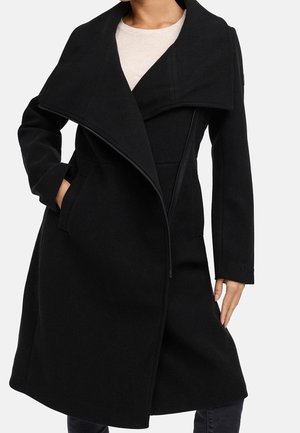 Woman wearing a knee-length black coat with wide lapels, side pockets, an asymmetrical front zipper, and a light beige top underneath.