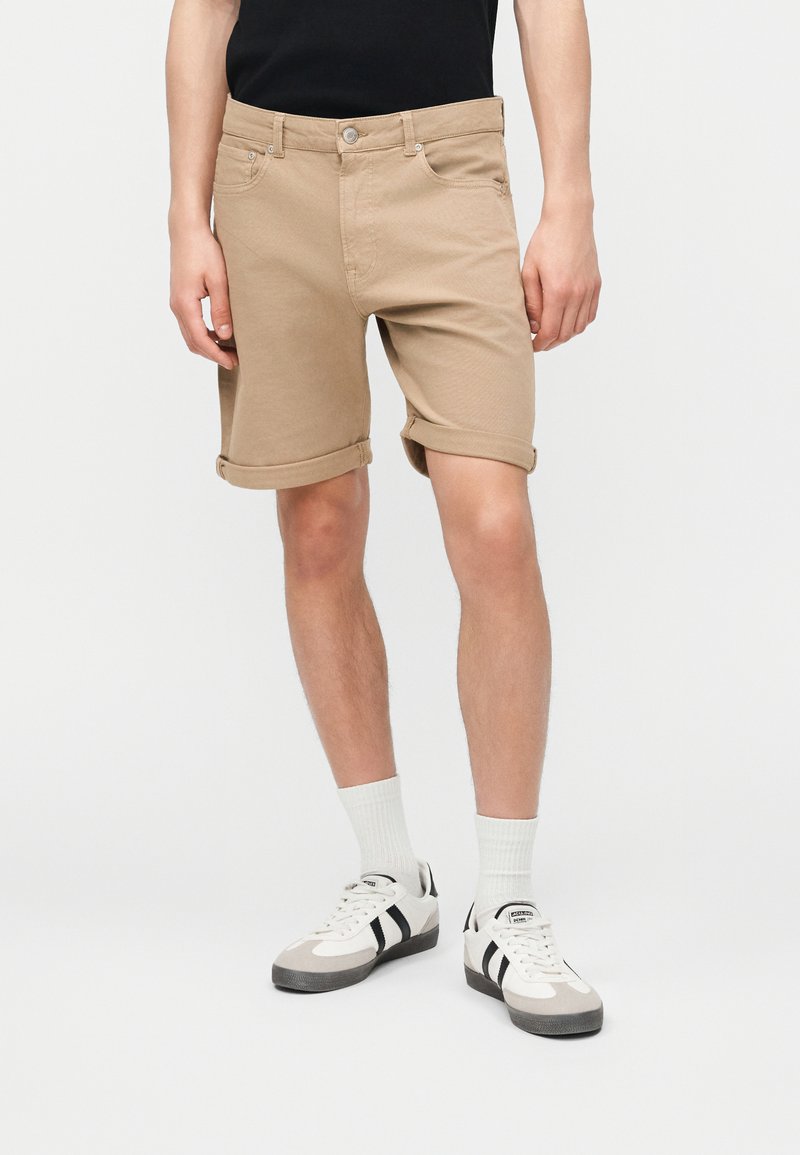 Homme portant un short beige retroussé, des chaussettes blanches, des baskets noires et blanches, et une chemise noire, debout devant un fond uni.