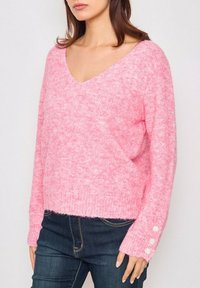 Pull en rose, duveteux avec un col en V, coupe ample, poignets côtelés ornés de trois boutonnières. Le matériau semble doux et texturé.