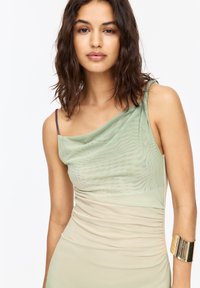 Elastisches, schichtweise gestaltetes Kleid mit einem asymmetrischen Design in Grün und Beige. Es hat einen drapierten Ausschnitt und ein goldenes Armreif.