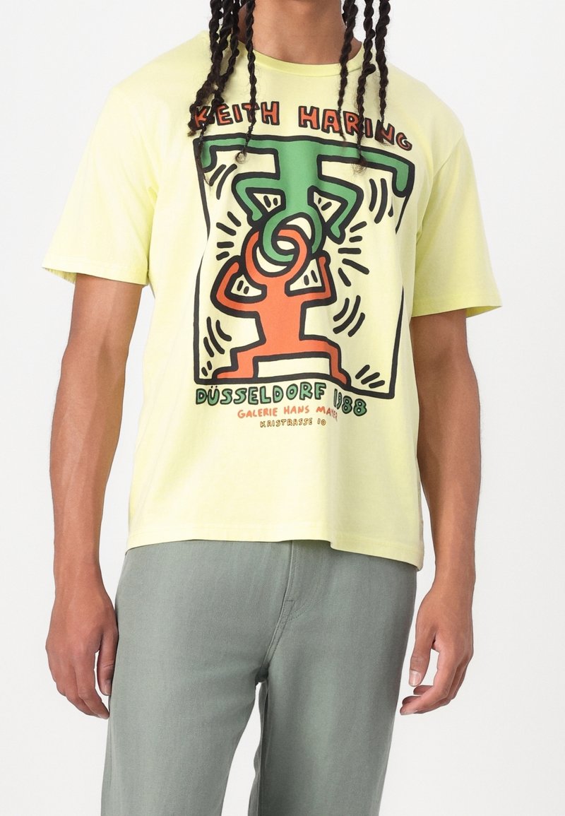 Personne portant un T-shirt jaune avec une œuvre colorée de Keith Haring représentant deux figures en contour et le texte "Düsseldorf 1988" sur le devant.