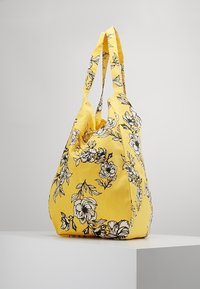 Borsa a mano gialla con motivo floreale nero, in tessuto morbido, design ampio e due robuste maniglie per il trasporto.