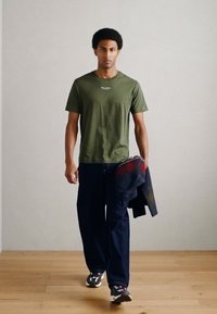 Λαδί κοντομάνικο μπλουζάκι, μπλε τζιν παντελόνι και πουλόβερ με σχέδια. Το μοντέλο φορά σκούρα αθλητικά παπούτσια και στέκεται σε ξύλινο πάτωμα.