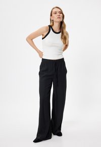 Schwarze weit geschnittene Hose mit elastischem Bund, kombiniert mit einem weißen, gerippten Tanktop mit einem schwarzen Kontrast-Ausschnitt. Glatte Stofftextur.