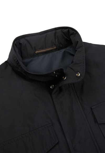 Czarna kurtka z wysokim kołnierzem, zapinana na zatrzaski, z przednimi kieszeniami z patkami, przedstawiona częściowo z widoczną podszewką wewnętrzną.