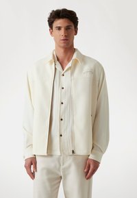 Cream-colored zip-up jacket over a textured beige shirt with brown buttons, worn with matching light-colored pants.