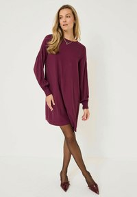 Donna che indossa un vestito bordeaux largo con maniche lunghe, collant neri trasparenti e décolleté bordeaux a punta, in piedi di fronte a uno sfondo bianco uniforme.