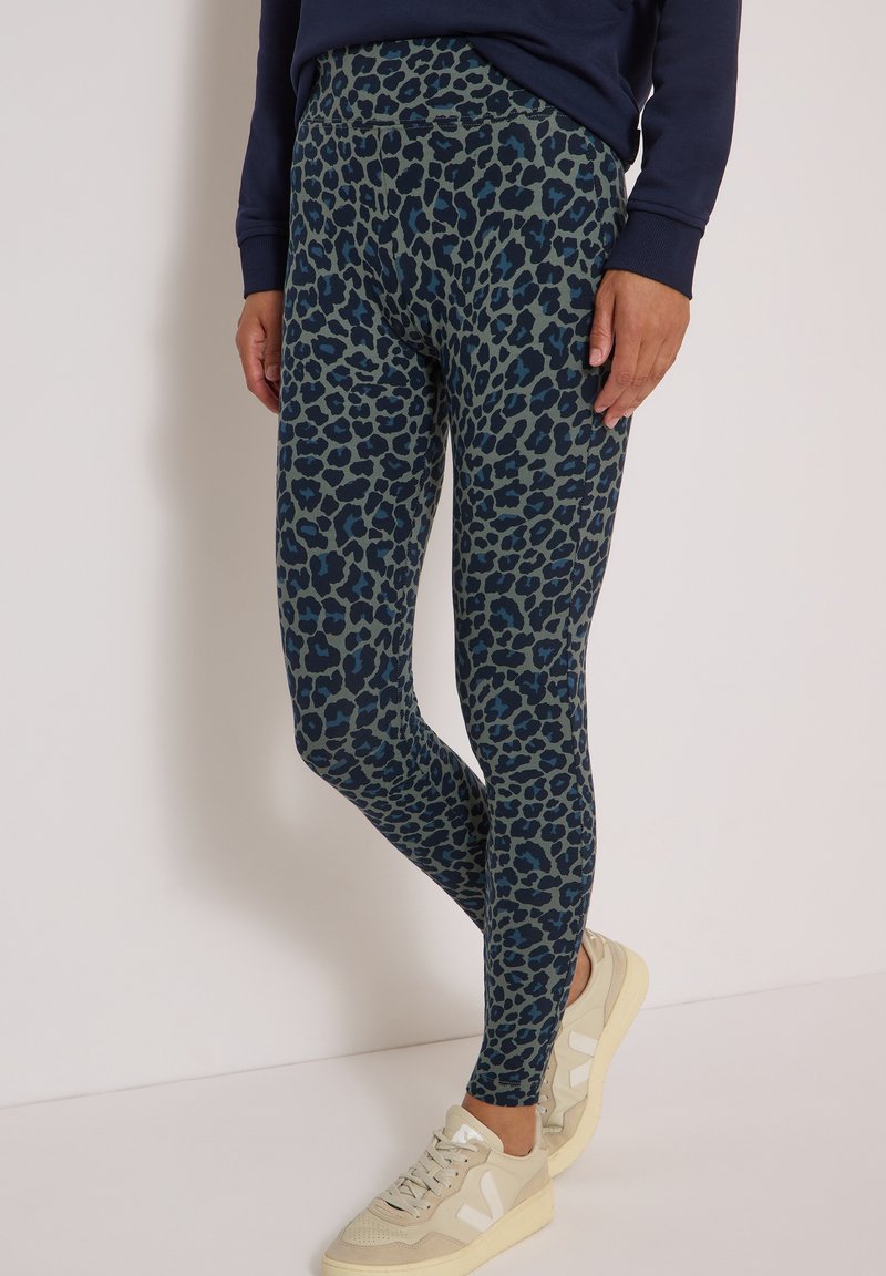 Personne portant des leggings bleu foncé et noir à imprimé léopard avec des baskets beiges sur un fond clair uni.