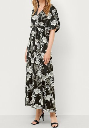 Mujer con vestido maxi negro con estampado floral blanco, escote en V, mangas cortas y anchas, y sandalias negras de tacón alto, de pie frente a una pared lisa.