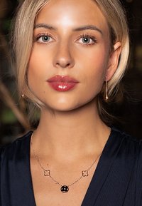 Gold necklace with three links: two floral shapes and a round pendant. The model wears a dark blue outfit and gold drop earrings.