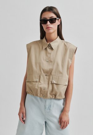 Top senza maniche di colore beige con colletto, due tasche frontali e fondo arricciato; indossato sopra pantaloni larghi blu chiaro.