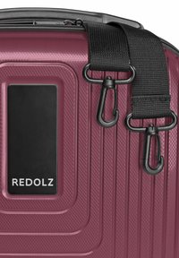 Close-up of burgundy hard-shell suitcase with textured surface, black detachable shoulder strap, and rectangular black label reading "REDOLZ."