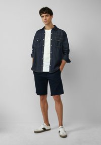 Jeanshemd met twee borstzakken, wit t-shirt eronder, marineblauwe shorts en casual sneakers. Natuurlijke textuur, ontspannen pasvorm en eenvoudig ontwerp.