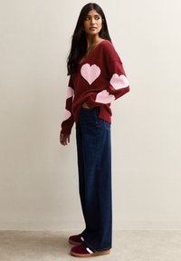 Maroon knitted sweater with pink heart patterns on sleeves, paired with wide blue denim jeans and burgundy sneakers with white accents.