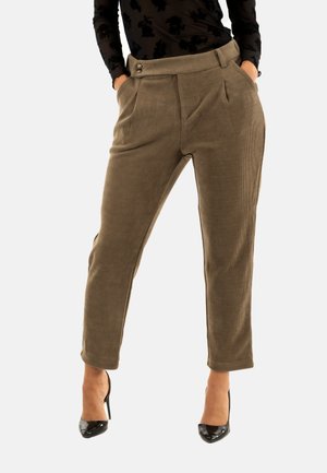 Donna che indossa pantaloni in velluto a coste marroni a vita alta e gamba affusolata, con tasche laterali e tacchi alti neri, con le mani in tasca, in piedi contro uno sfondo bianco.